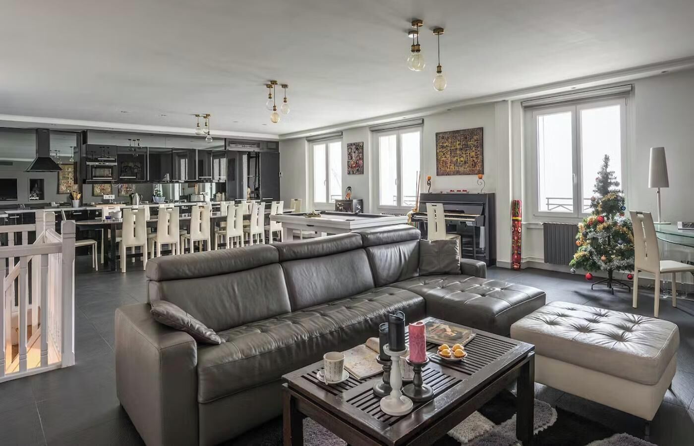 A modern living room with a grey leather sofa, a coffee table with decorative items, a dining area with white chairs, and a piano.