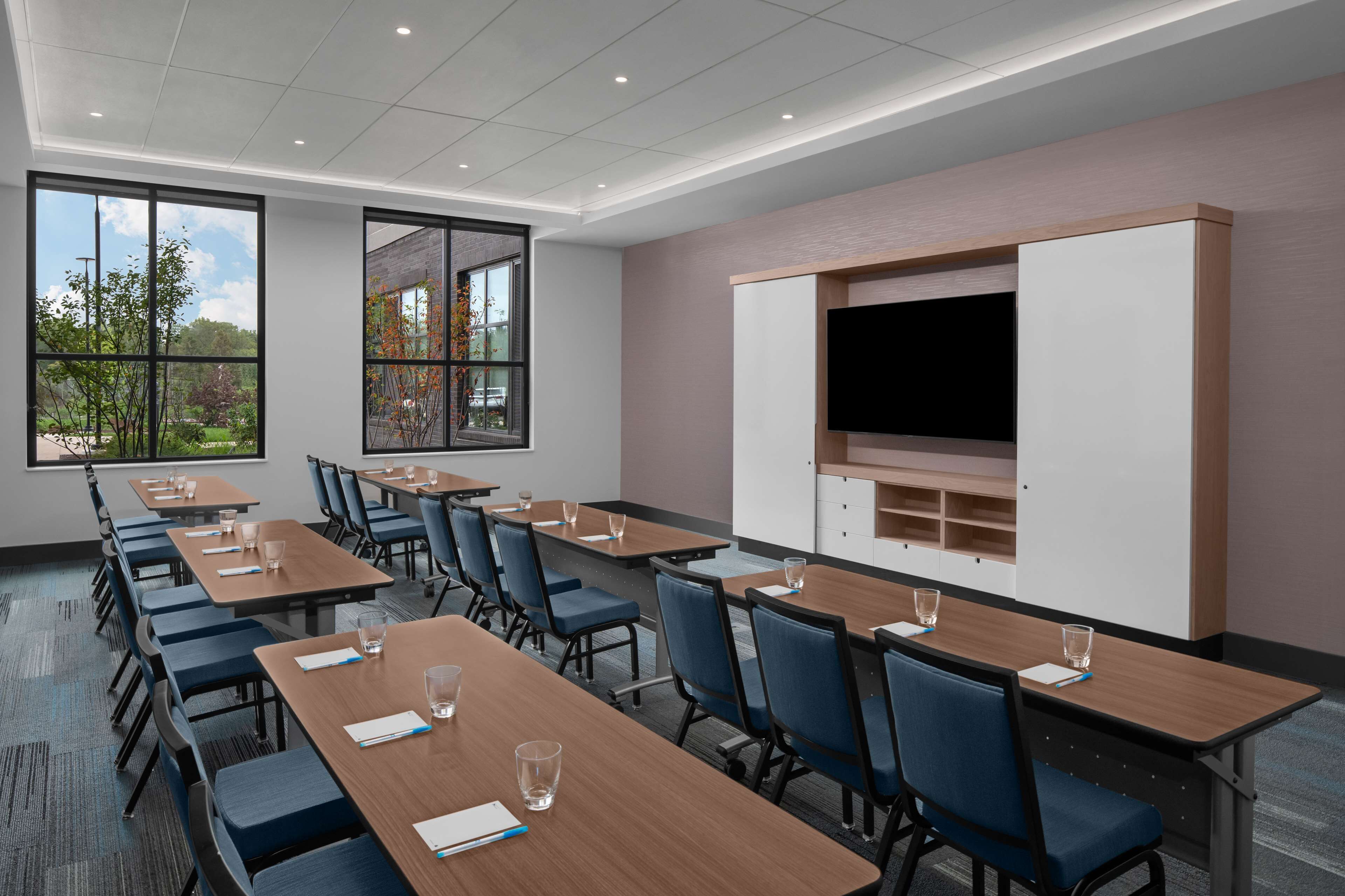 A conference room with multiple tables, chairs, a large screen, and windows offering a view of trees and a clear sky.