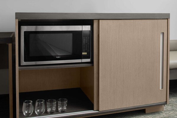 Una kitchenette compacta con microondas, un estante con vasos y un armario de madera.
