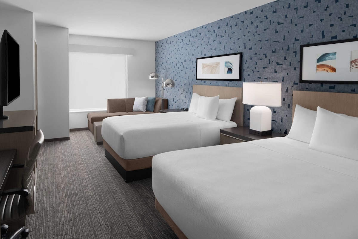 Ein Hotelzimmer mit zwei Betten, einem Schreibtisch, einem Fernseher und einem Fenster.