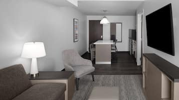 A modern hotel room with a grey sofa, a small table with a lamp, a grey armchair, a kitchenette with a bar counter, and a flat-screen TV mounted on the wall.