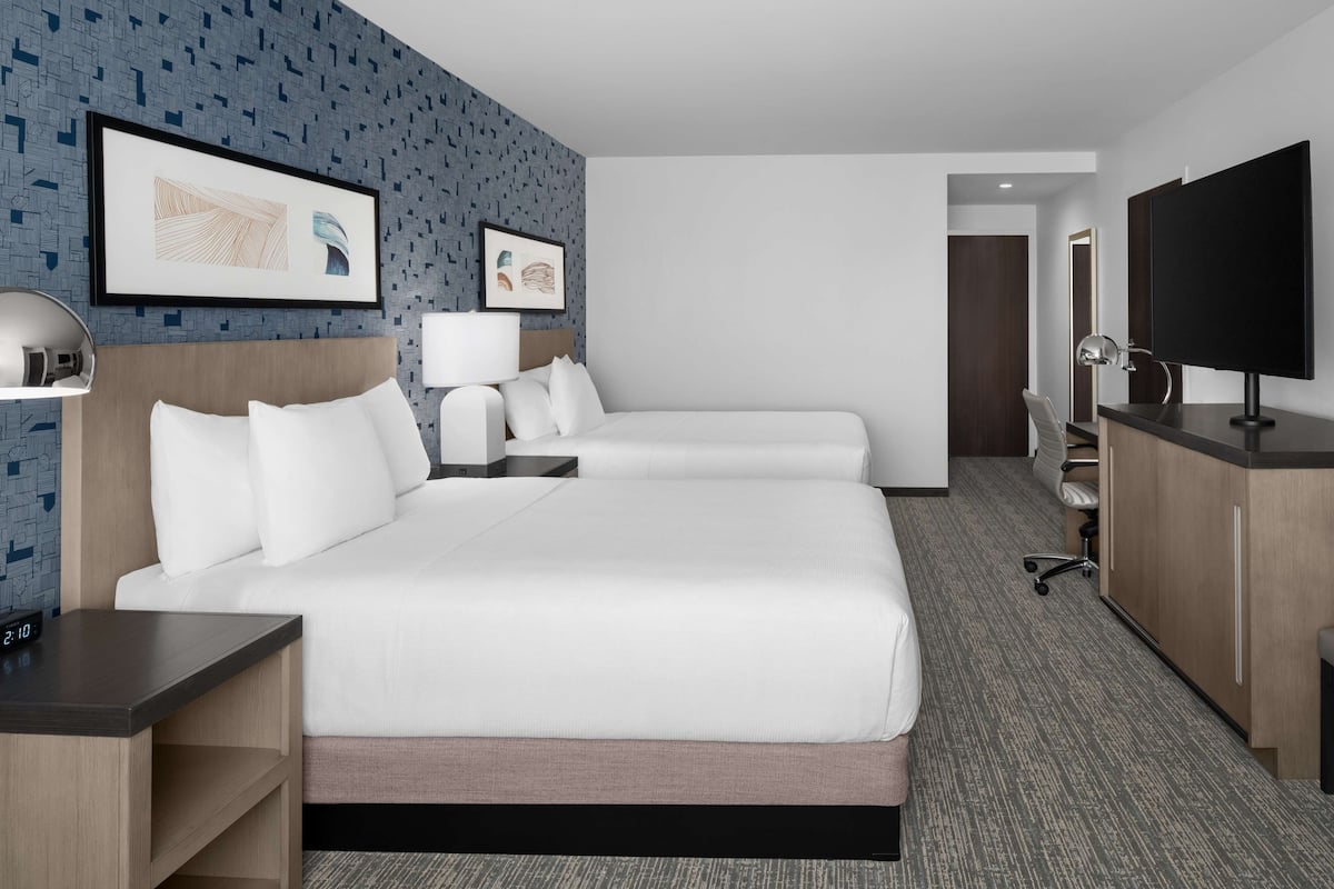 Ein Hotelzimmer mit zwei Betten, einem Schreibtisch, einem Sessel, einem Fernseher und Bildern an den Wänden.
