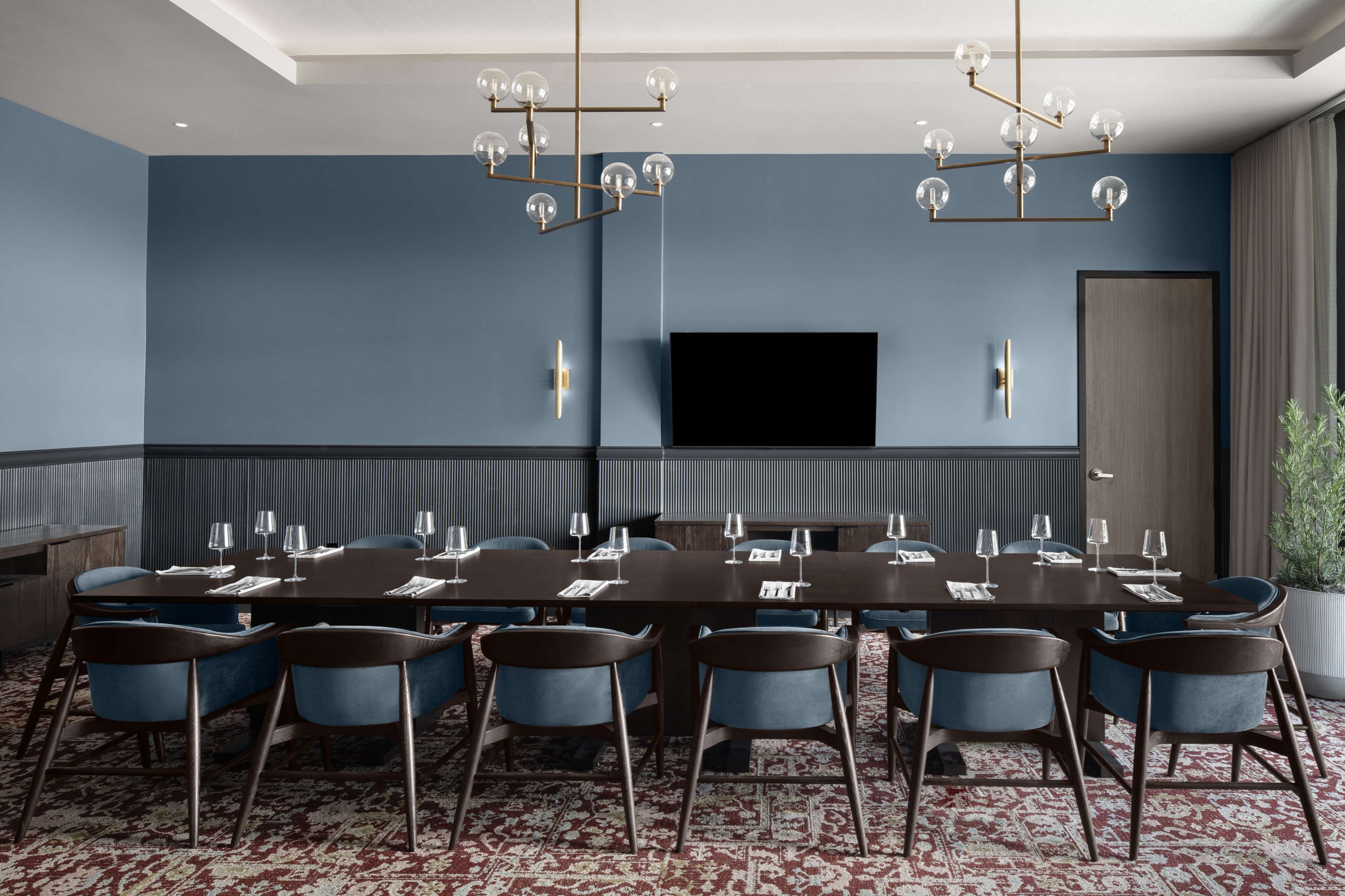 A conference room with a long table, blue chairs, a chandelier, a flat-screen TV, and a potted plant.