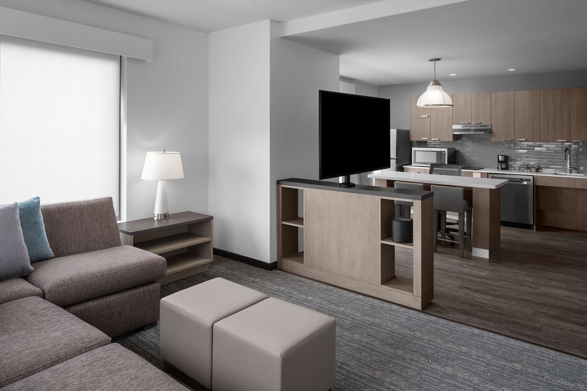 Ein modernes Wohnzimmer mit einer Couch, einem Hocker und einem Fernseher. Es gibt eine Küchenzeile mit einem Esstisch und Stühlen.