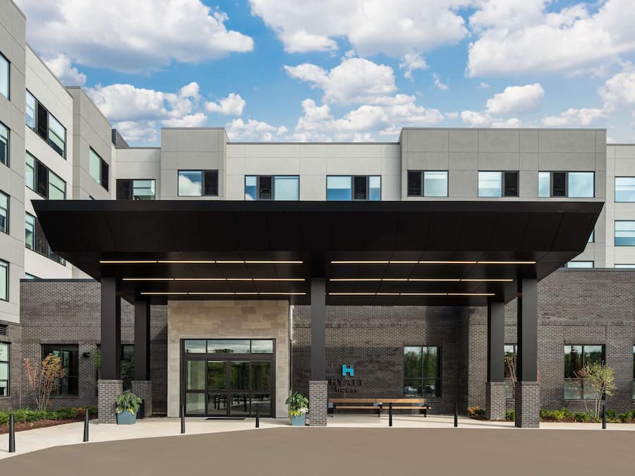 Entrada moderna de hotel com área de espera coberta, janelas amplas e placa 'Hyatt House' visível.