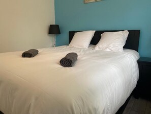 A neatly made bed with white linens and two rolled towels, a black nightstand with a lamp, and a teal wall. - Petit Gîte en Auvergne Entièrement Rénové (Giat)