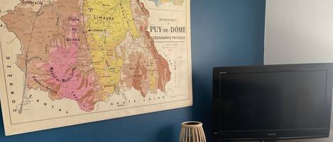 Une pièce avec une grande carte ancienne accrochée au mur, un téléviseur à écran plat posé sur un meuble, une armoire blanche avec des tiroirs et une étagère remplie de livres et d’un haut-parleur.