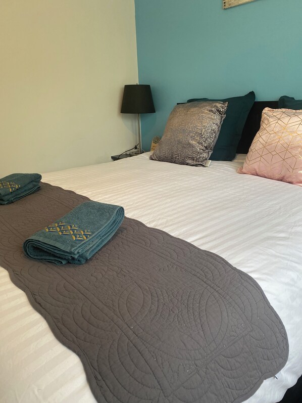 A neatly made bed with a brown quilt and two folded towels, a lamp, and patterned pillows on a blue wall. - Petit Gîte en Auvergne Entièrement Rénové (Giat)