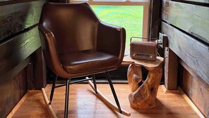 A cozy cabin interior with a wooden rocking chair, a wooden side table with a radio, and a large window offering a view of the outdoors. - Remodeled Cook Forest Cabin (Clarington)