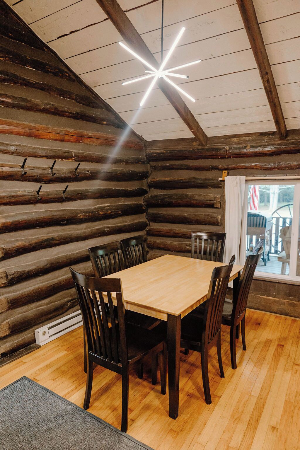 A log cabin interior with a wooden dining table and chairs, a star-shaped ceiling light, and a view of the outside through a window.