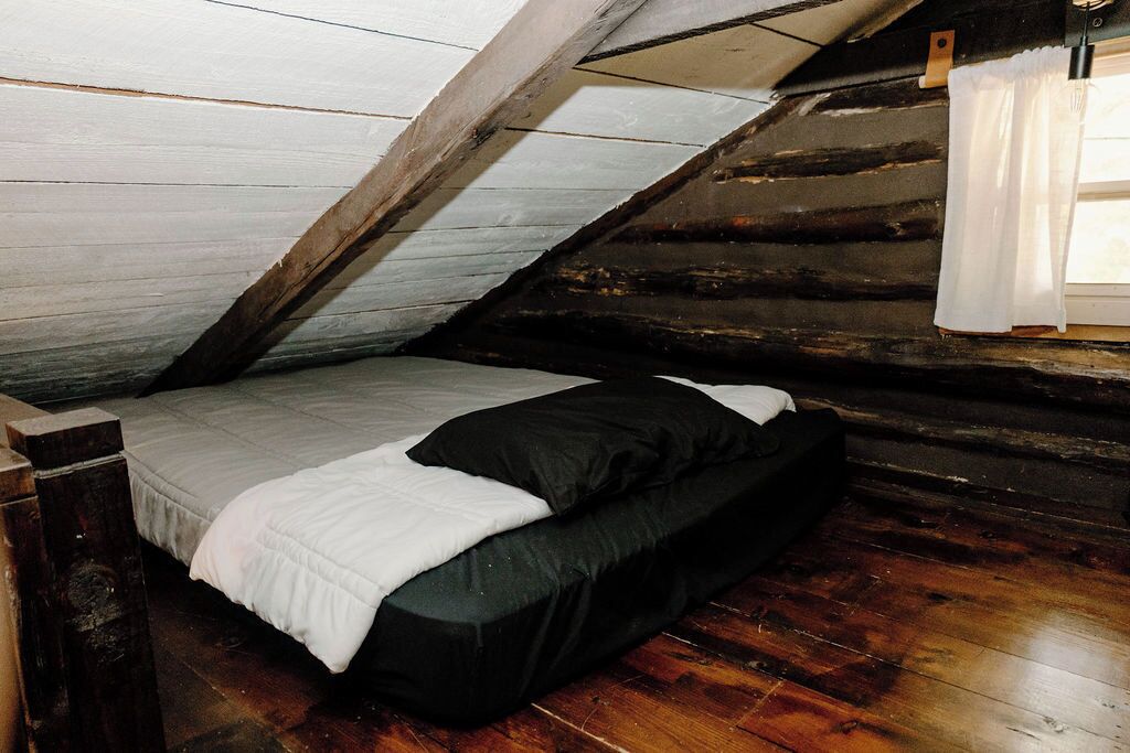 A small, cozy attic bedroom with a wooden floor, a single bed with white and black bedding, and a window with a white curtain.