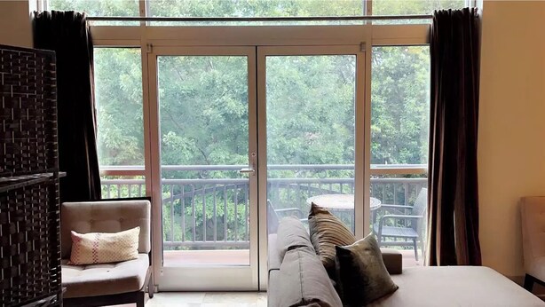 A room with a large sliding glass door leading to a balcony with a view of trees. There is a bed with pillows and a chair with a cushion. - Lovely Condo in the Very Heart of NoDA (Charlotte)