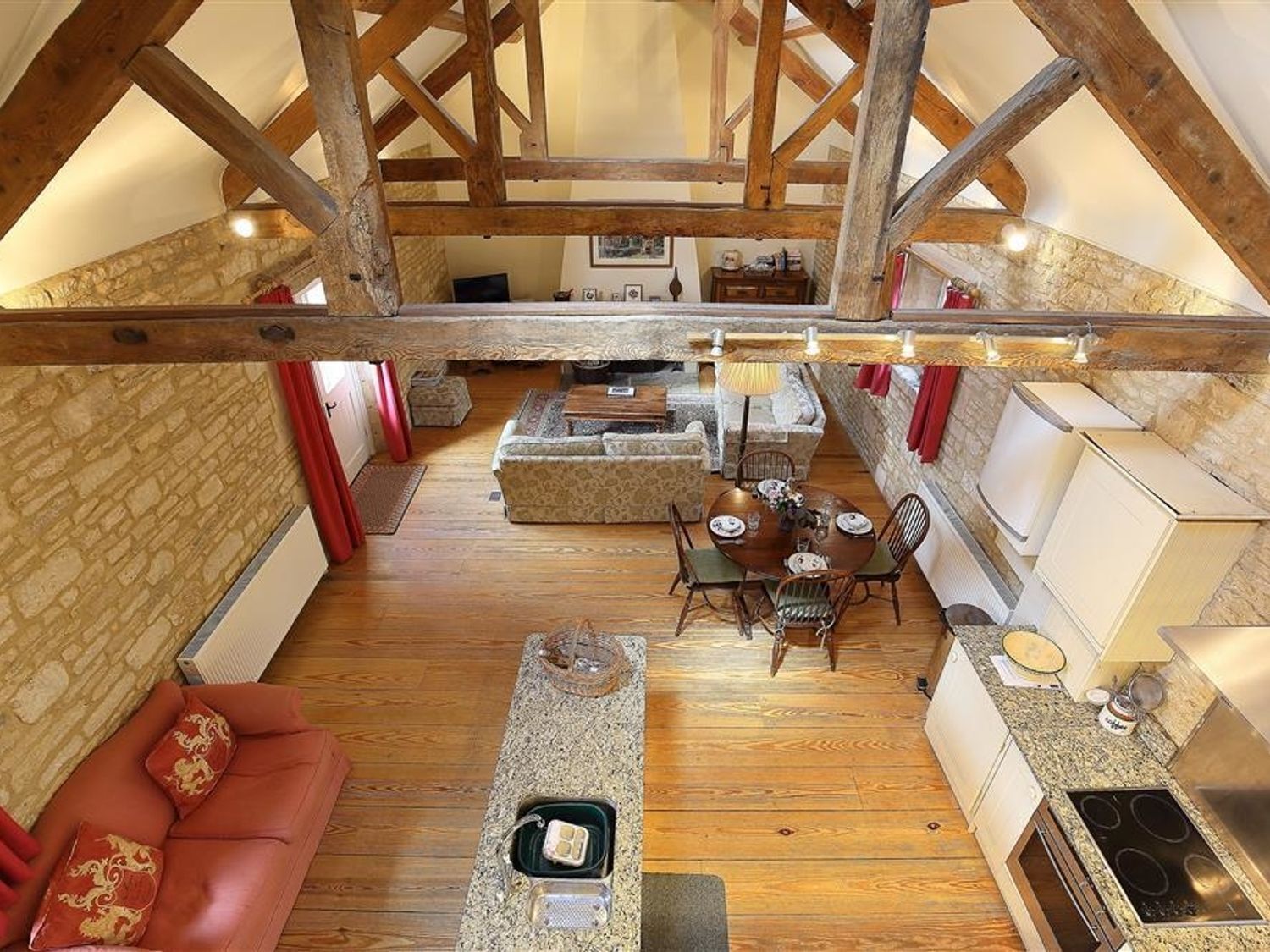 A spacious loft with exposed wooden beams, a stone wall, a cozy red sofa, a dining area with a table set for four, and a kitchenette with a stove and sink.