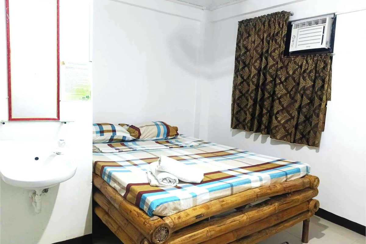 A small, simple room with a wooden bed, a white sink, and a window with patterned curtains.