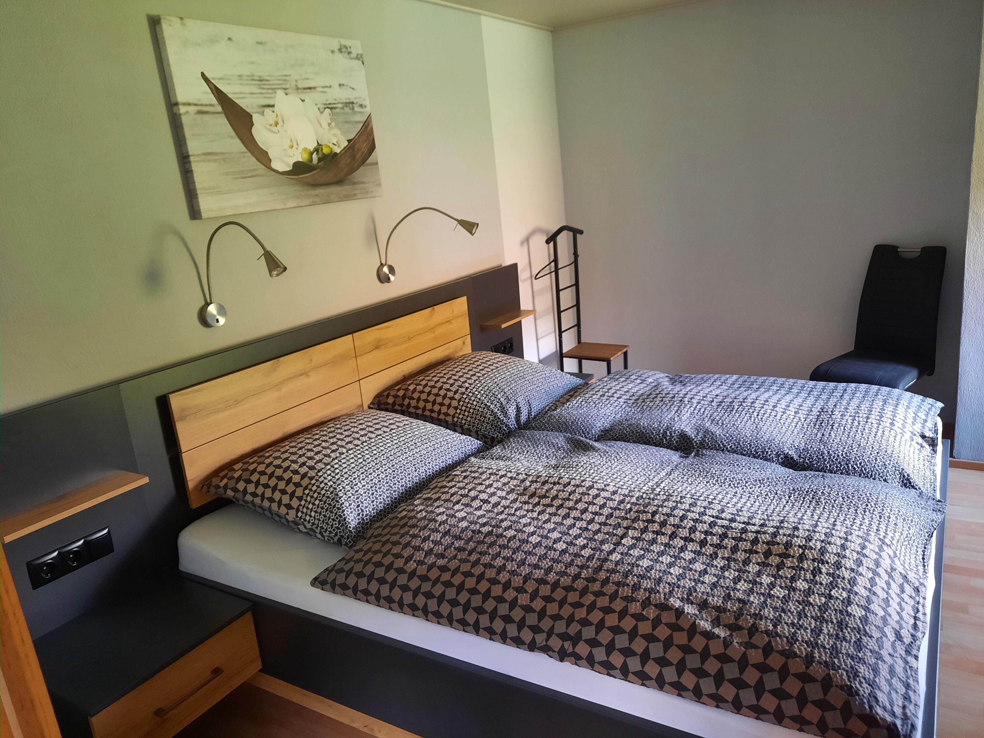 A double bed with patterned bedding, a bedside table, a wall-mounted lamp, and a small stool.