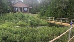 Eine Holzterrasse mit Seilbarriere und Metallpfählen, umgeben von dichtem Grün und einem Gebäude im Hintergrund.