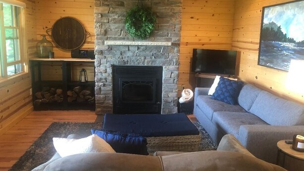 Ein gemütliches Blockhauss-Interieur mit Kamin aus Stein, einem grauen Sofa, einem Fernseher und einem großen Fenster.