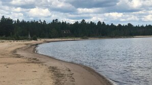 Ein Sandstrand mit einem ruhigen See, umgeben von dichtem Wald und einigen Gebäuden in der Ferne.