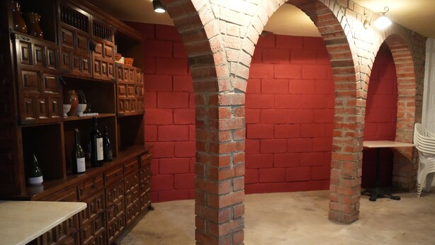 A room with a brick fireplace, wooden shelving with wine bottles, and a white chair. - Casa Rural Margarita (Peñaranda de Duero)