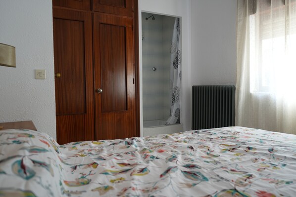 Une chambre avec une armoire en bois, une douche avec une porte en verre, un radiateur et un lit recouvert d’une couvre-lit à motifs floraux.