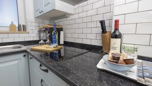 Une cuisine avec des meubles blancs, un plan de travail noir, une planche à découper en bois, des verres à vin, une bouteille de vin et un bol de nourriture.