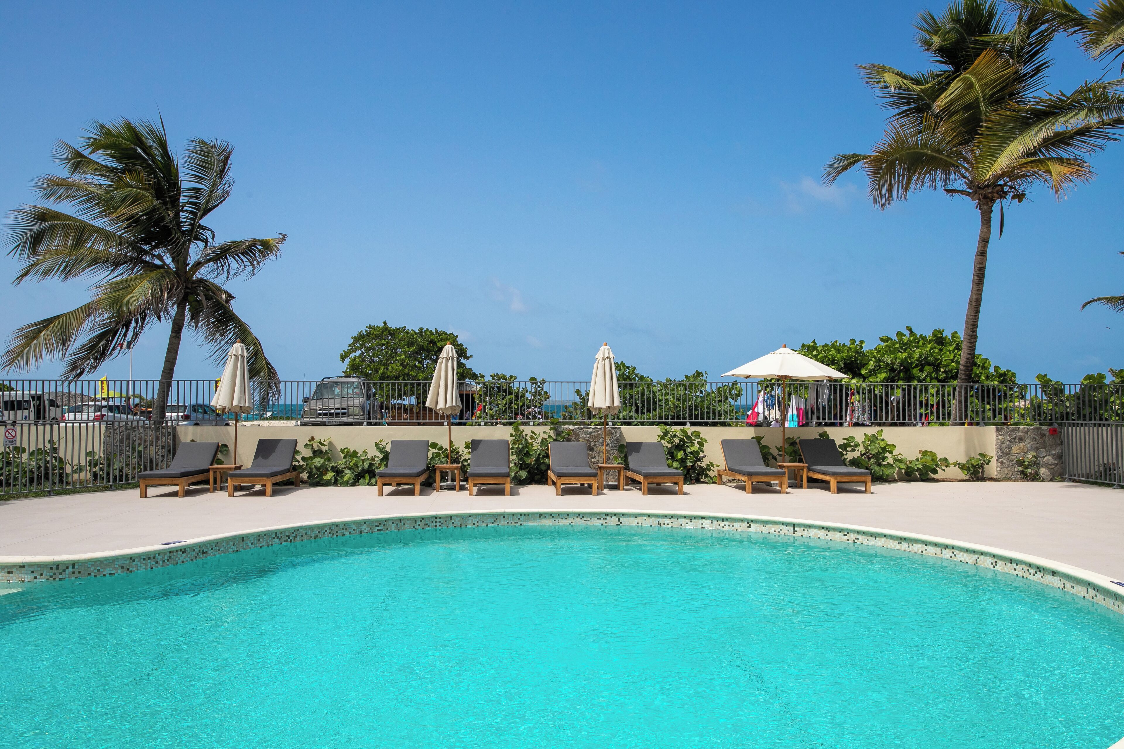 A pool area with lounge chairs and umbrellas, surrounded by palm trees and a clear sky.