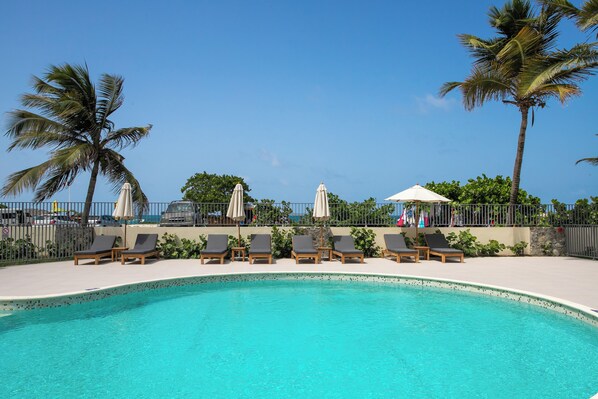 A pool area with lounge chairs and umbrellas, surrounded by palm trees and a clear sky. - Appartement vue Mer, Accès Direct Plage D'orient Bay (Orient-Bay)