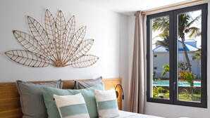 A bedroom with a bed, pillows, a wall art piece, and a window with a view of a pool and palm trees. - Appartement vue Mer, Accès Direct Plage D'orient Bay (Orient-Bay)