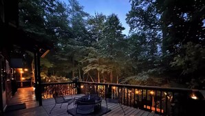 Une terrasse en bois avec un foyer et des chaises, entourée d’arbres et d’une rambarde.