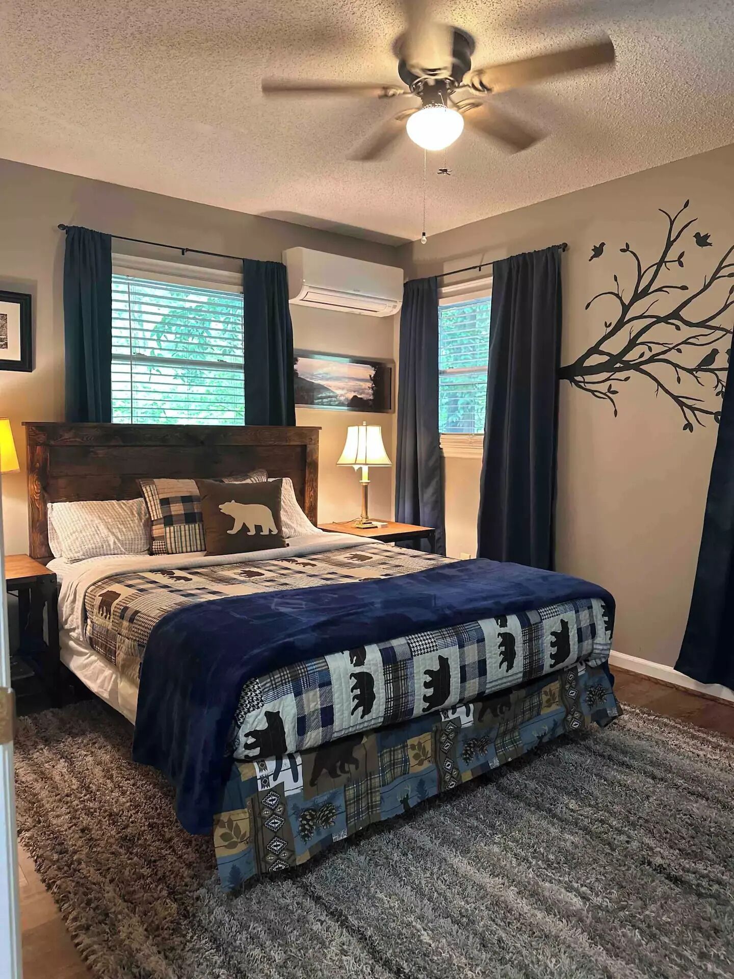 A bedroom with a bed, a ceiling fan, a nightstand with a lamp, and a wall-mounted air conditioner.