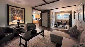 Un salon comprenant un canapé, une table basse et un fauteuil. On aperçoit également un coin repas avec une table et des chaises, visibles à travers une porte ouverte.