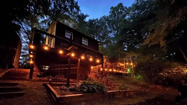 Un chalet en bois rustique, doté d’une terrasse et illuminé de guirlandes lumineuses, et entouré d’arbres.