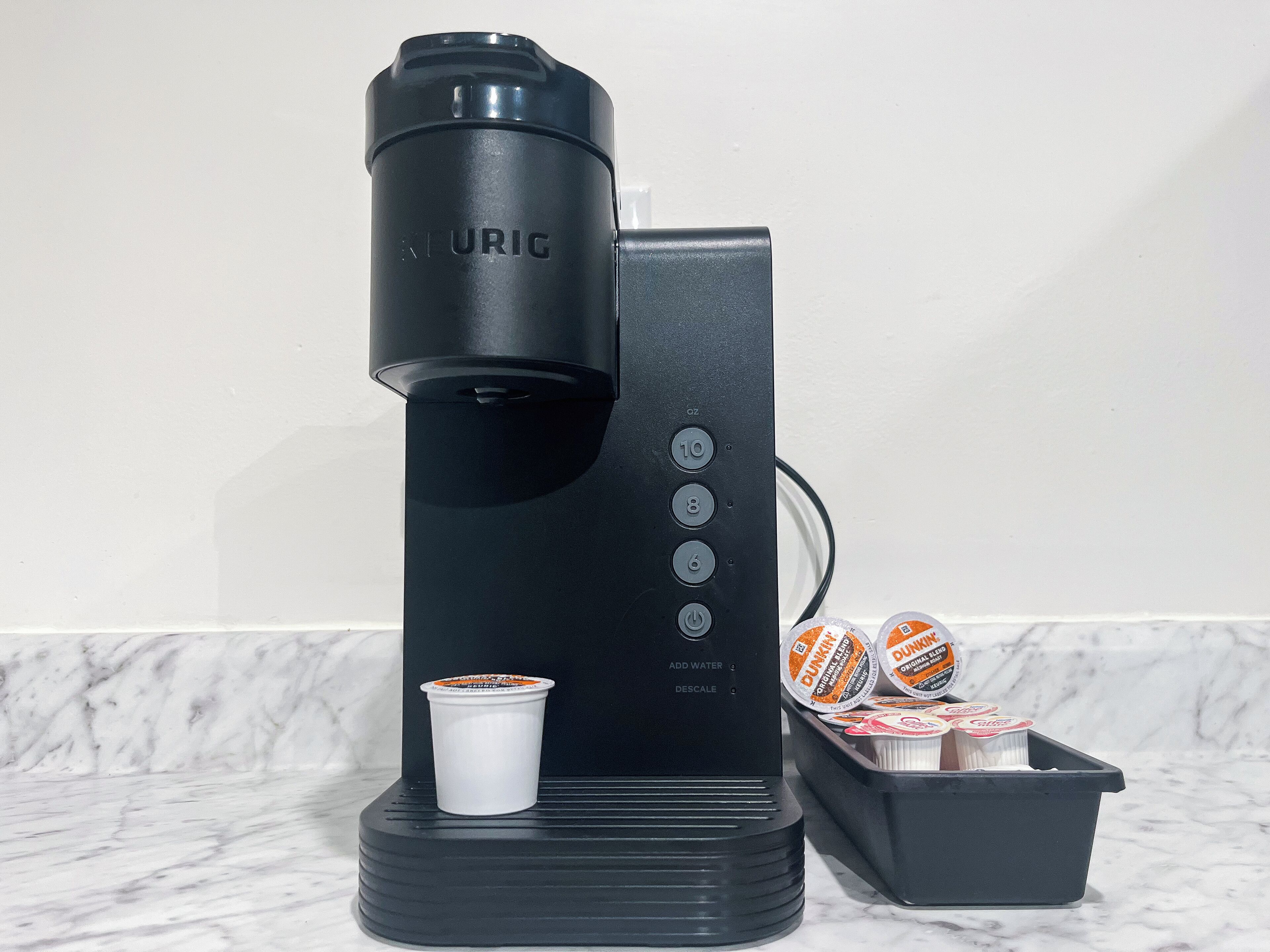 A Keurig coffee maker with a cup of coffee, a container of Dunkin' Donuts coffee pods, and a black tray.
