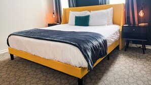 Un lit avec une tête de lit jaune, une literie blanche et un plaid foncé. Il y a deux tables de chevet avec des lampes, dont l’une est dotée d’un abat-jour décoratif. La pièce est agrémentée d’un tapis à motifs et d’une fenêtre avec des rideaux.
