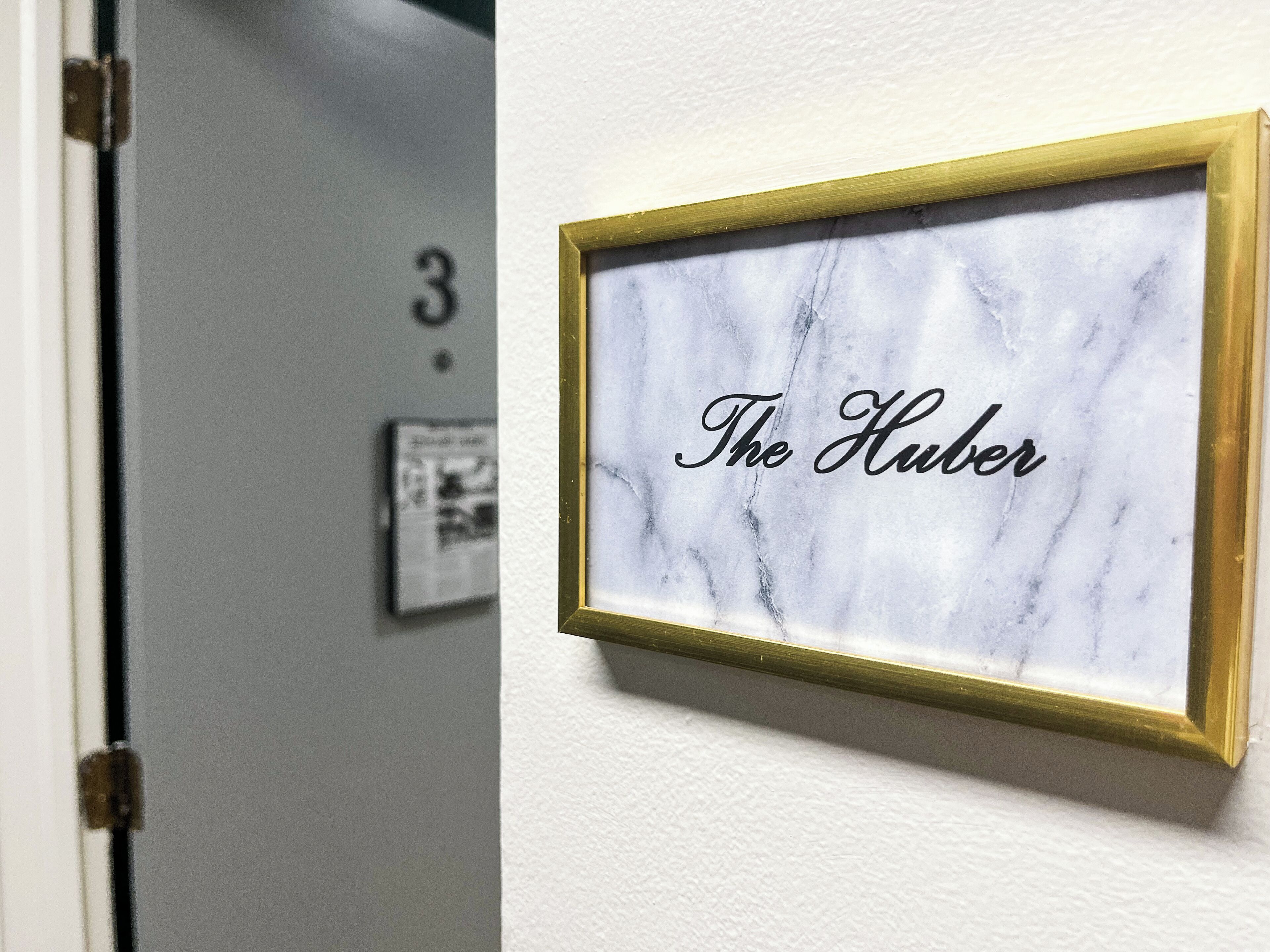 Un cartel enmarcado con la inscripción "The Huber" sobre una pared de mármol, una puerta con el número "3" y un pequeño cuadro.