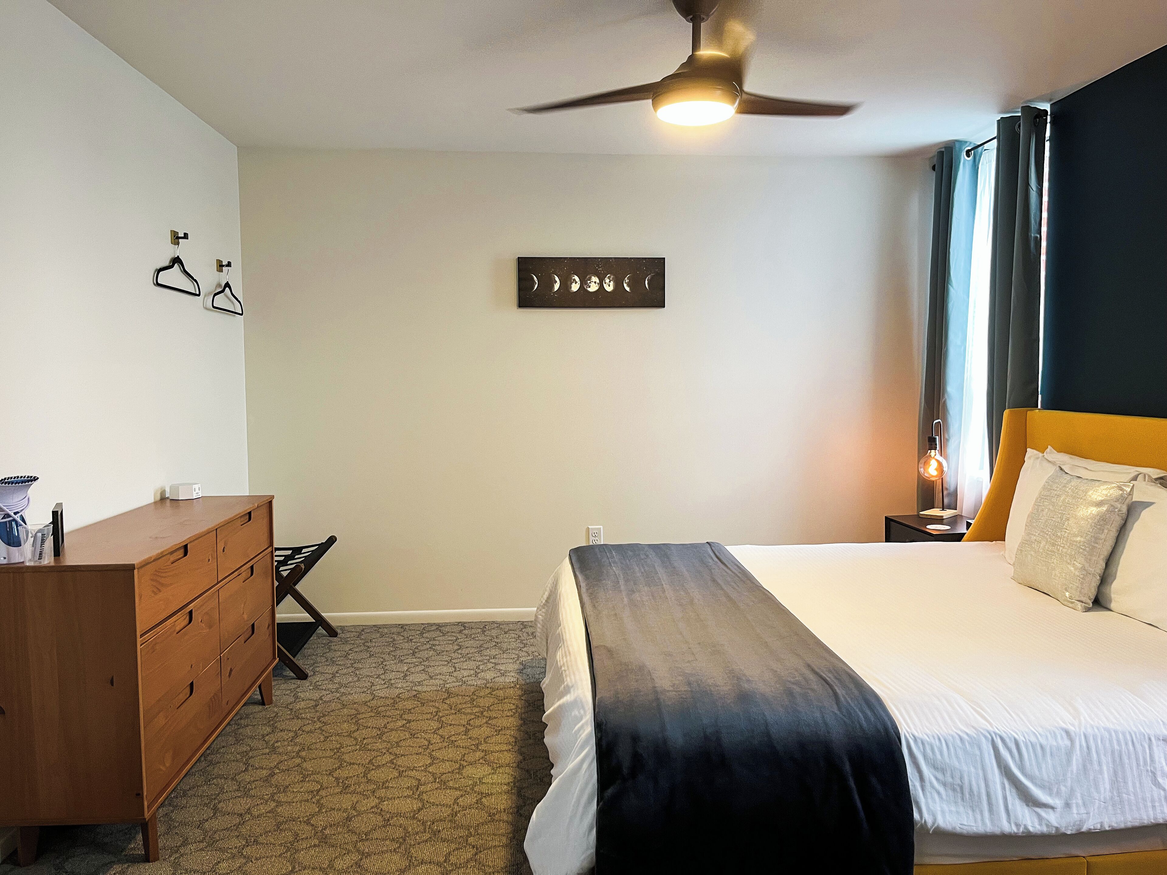 A hotel room with a bed, a wooden dresser, a ceiling fan, a wall-mounted artwork, and a nightstand with a lamp.