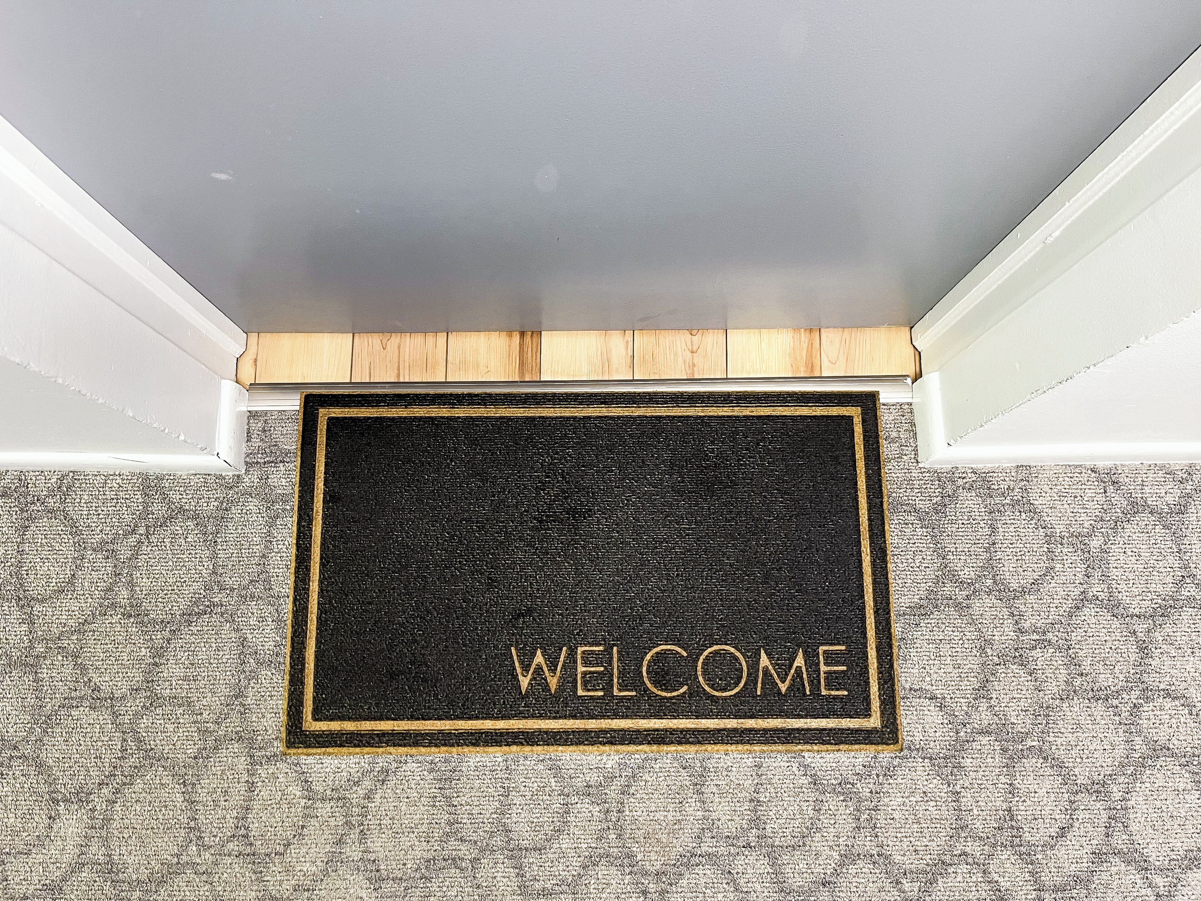 Tapete de boas-vindas com a palavra 'WELCOME' em letras douradas, colocado na entrada de um quarto com carpete texturizado e detalhes em madeira.