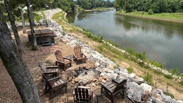 Een camping aan de rivier met een vuurkuil, houten stoelen en een tafel.