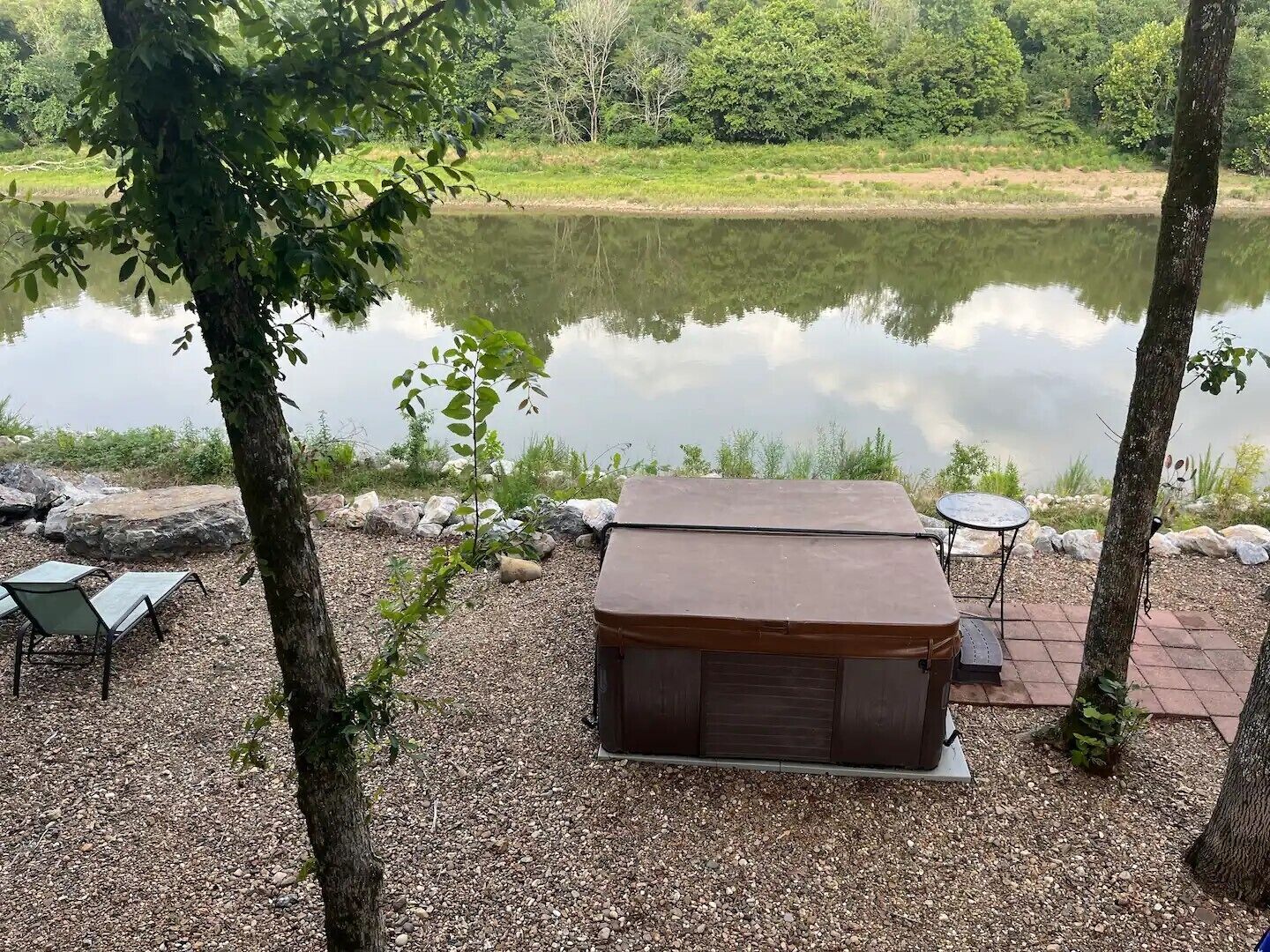 A hot tub with a wooden cover, surrounded by pebbles and trees, near a calm lake.