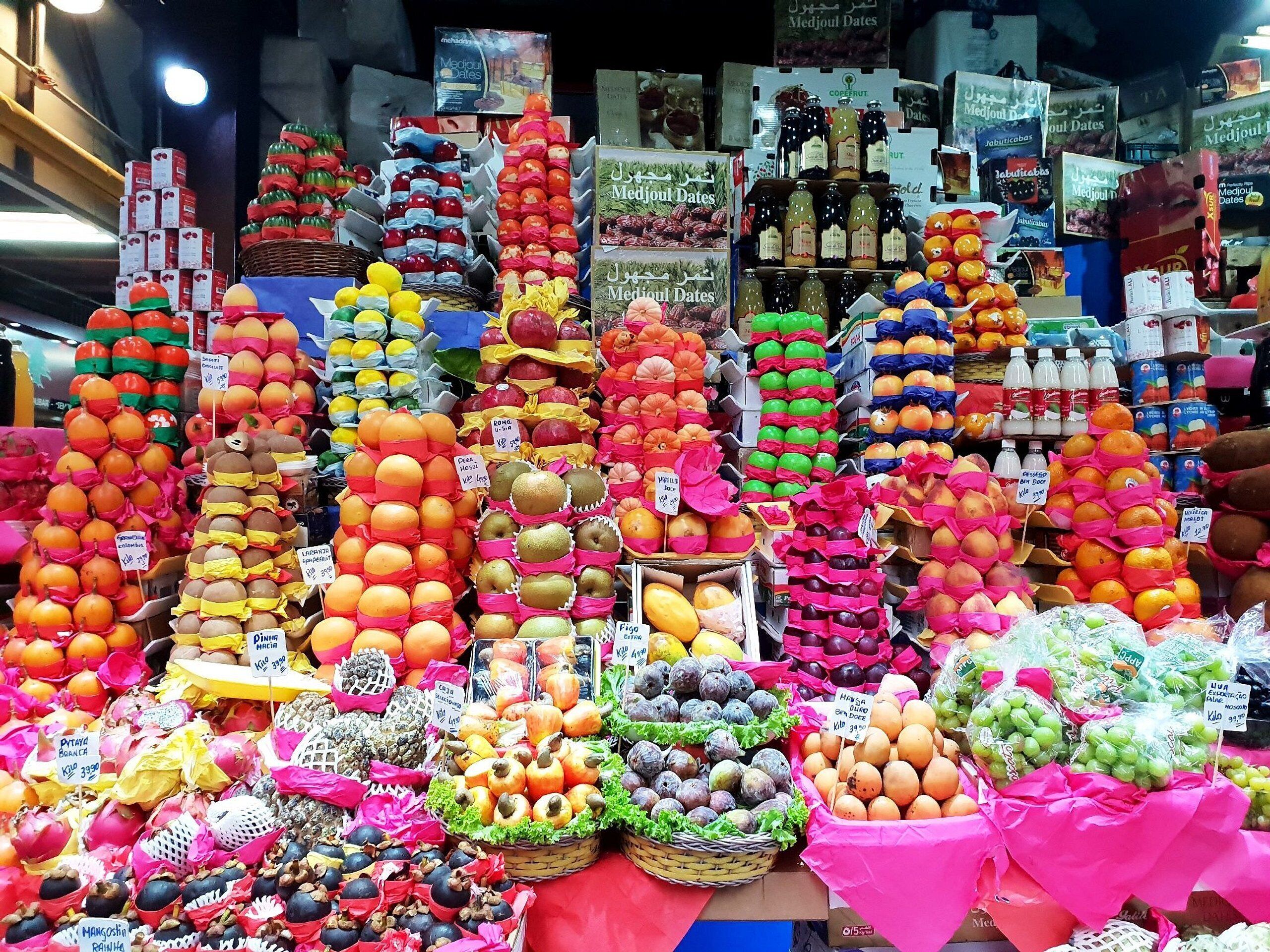 Un puesto de mercado con frutas y verduras dispuestas en cestas y estantes, con etiquetas de precio visibles.