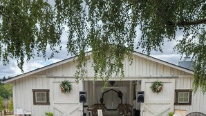 Un montaje para comedor al aire libre con una larga mesa de madera, centros de mesa florales y un edificio rústico de estilo granero al fondo.