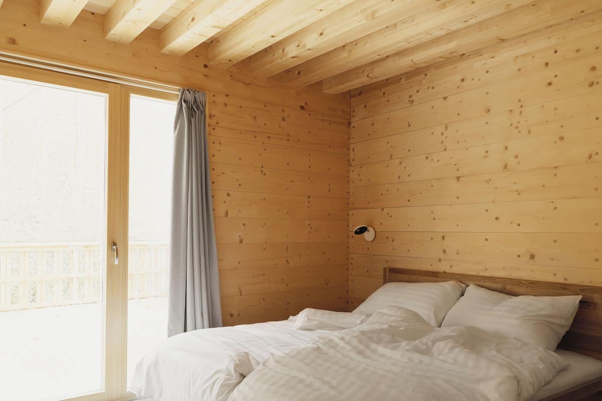 Ein Schlafzimmer mit Holzboden, einem Bett mit weißer Bettwäsche, einem Fenster mit Vorhang und Blick auf einen Balkon.