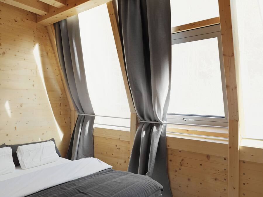 Une chambre avec un plafond en bois, un lit avec une literie blanche et grise, et une fenêtre avec des rideaux gris.