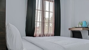 A neatly made bed with white linens, a window with sheer curtains, and a small table with a kettle and bottles. - Capital O 92583 Raja Soeta Hostel Syariah (Bandar Lampung)