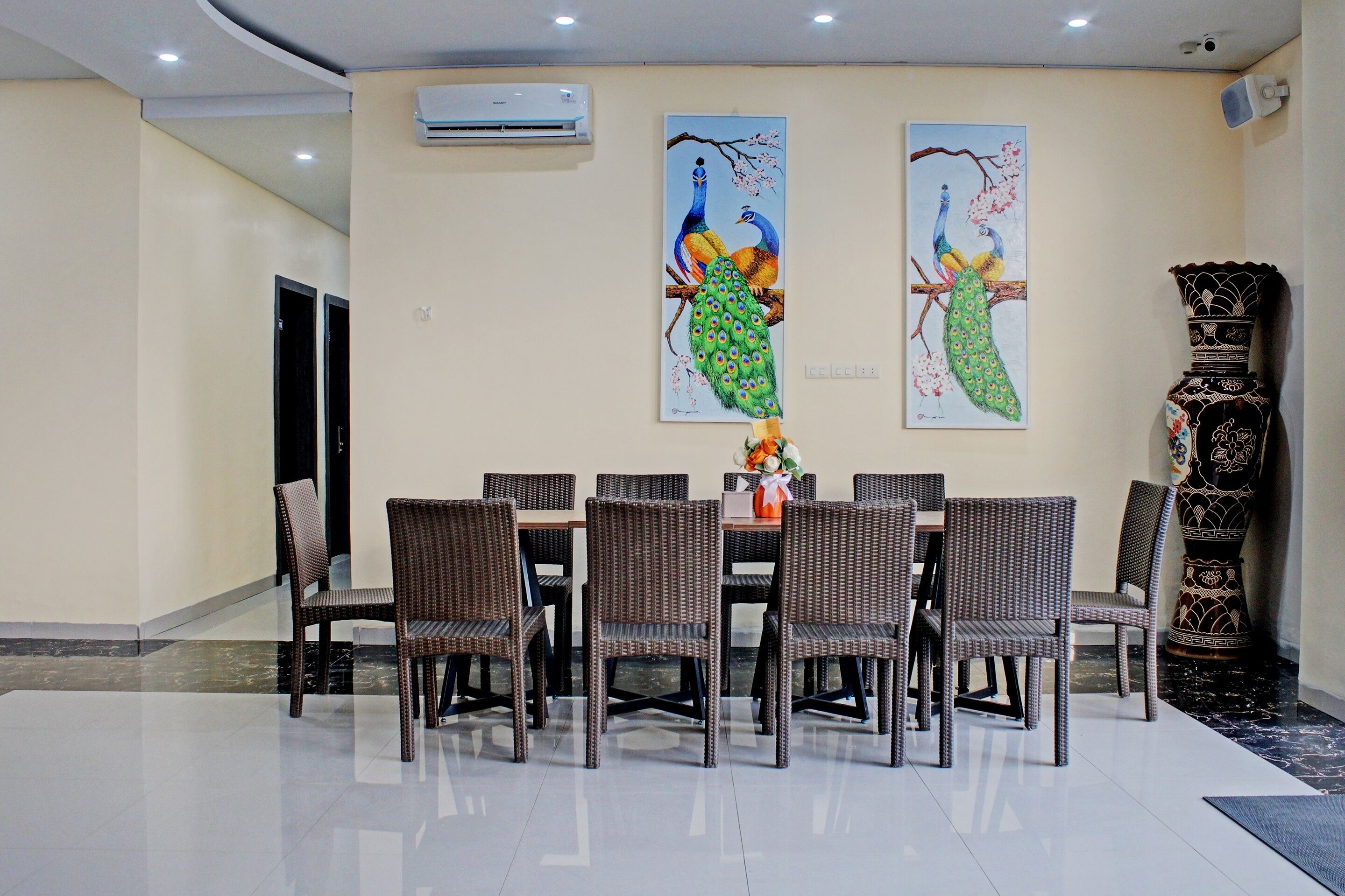 A dining area with a round table and eight wicker chairs, two wall-mounted paintings of peacocks, and a decorative black and white vase.