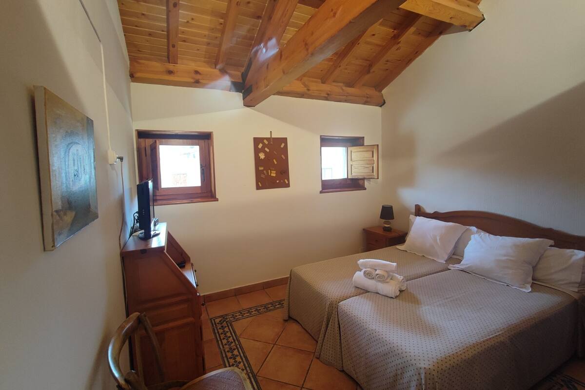 A bedroom with a wooden ceiling, two beds, a desk, a chair, and a small window.