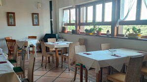 A dining area with multiple tables and chairs, large windows with curtains, and potted plants on the windowsill. - Posada Restaurante de Serrada (Puentes Viejas)