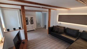 A modern living room with a wooden ceiling, a black sofa, a TV on a wooden stand, and a white cabinet with glass doors. - Holiday home in the middle of the old town of Goslar for up to 6 people (Goslar)