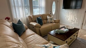 Una sala de estar con un sofá color beige, un sillón de cuero marrón, una mesa de centro de madera y un televisor de pantalla plana montado en la pared.