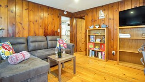 Een knusse woonkamer met een houten lambrisering, een grijze bank met bloemenkussens, een klein houten tafeltje met een fles en glazen, en een boekenkast met verschillende boeken en decoratieve objecten.